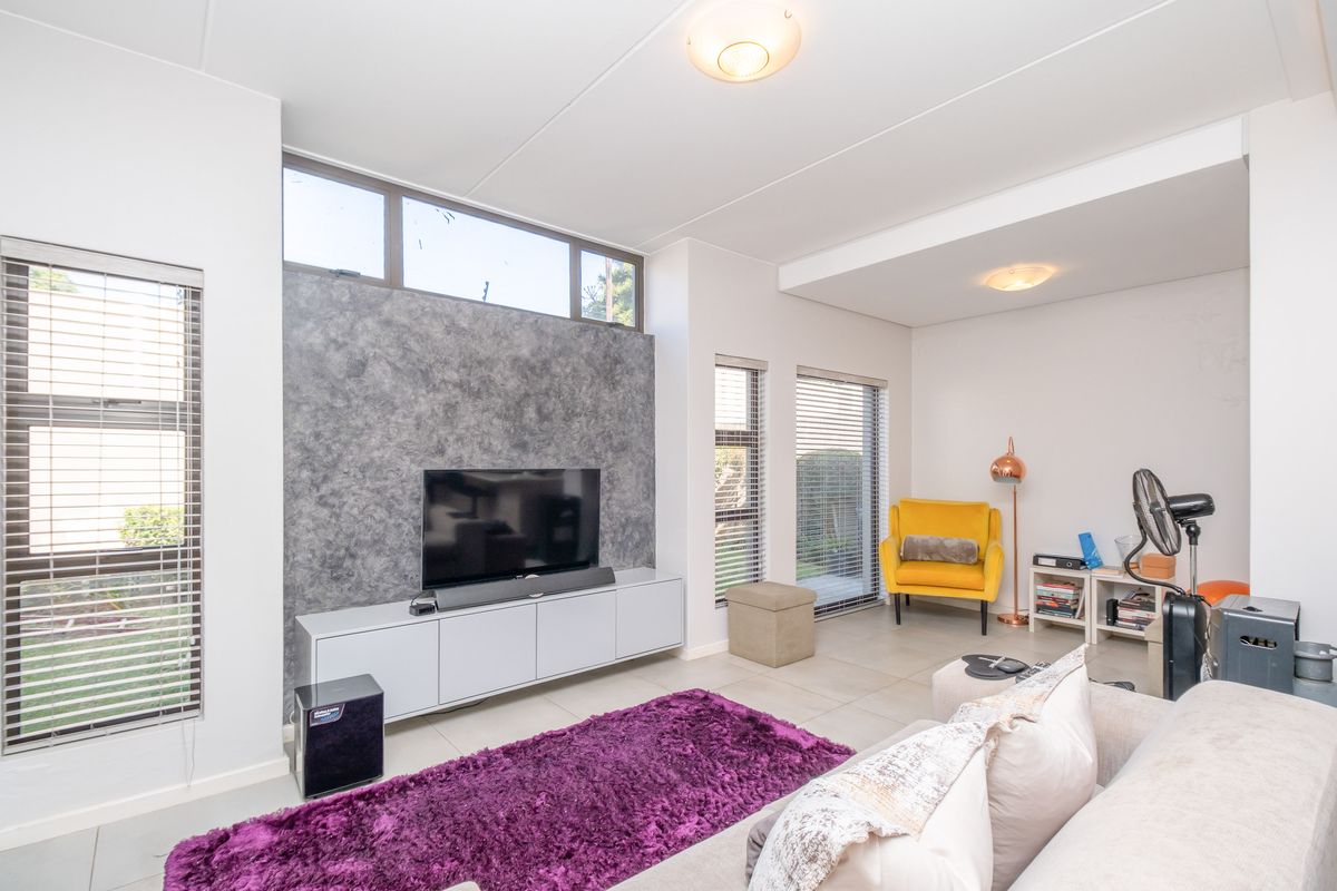 A bright living room featuring a textured feature wall, built-in media cabinetry, tiled flooring, and large windows with venetian blinds.