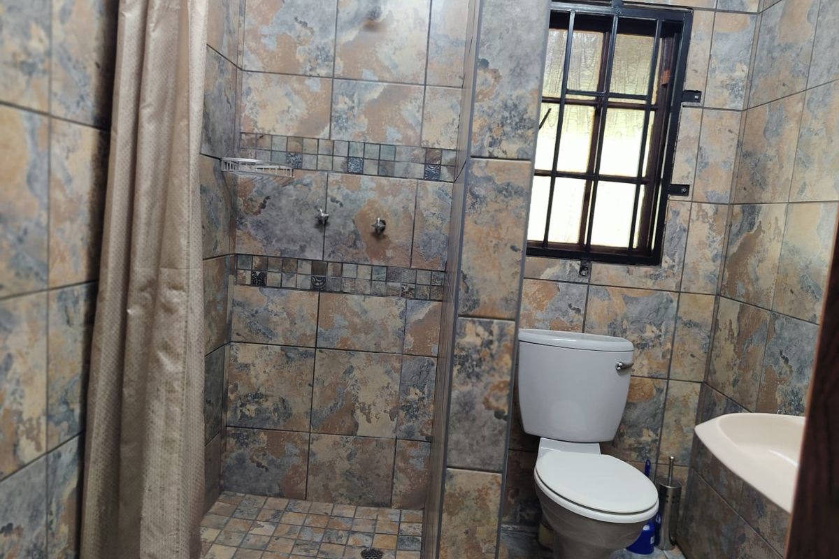 A functional bathroom featuring full-height stone-look wall tiling, a walk-in shower area, a standard toilet, and a small window with a security grille.