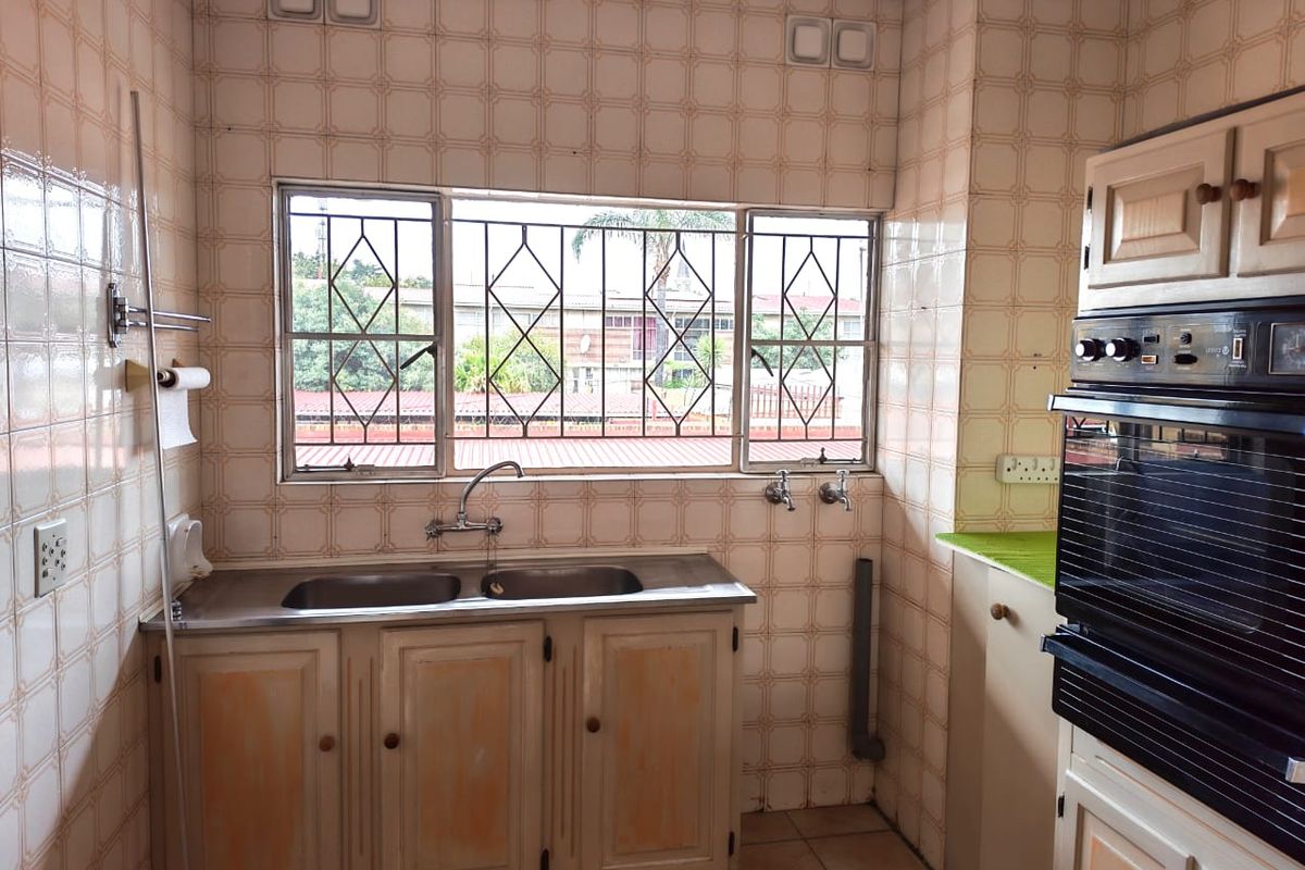 A dated kitchen featuring a double stainless steel sink, built-in oven, and extensive wall tiling with security bars on the window.
