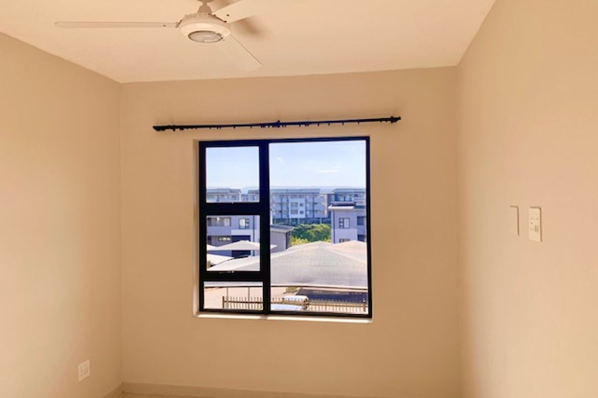 A bright, empty bedroom featuring neutral walls, tiled flooring, a ceiling fan, and a window with a view of the surrounding apartment complex.