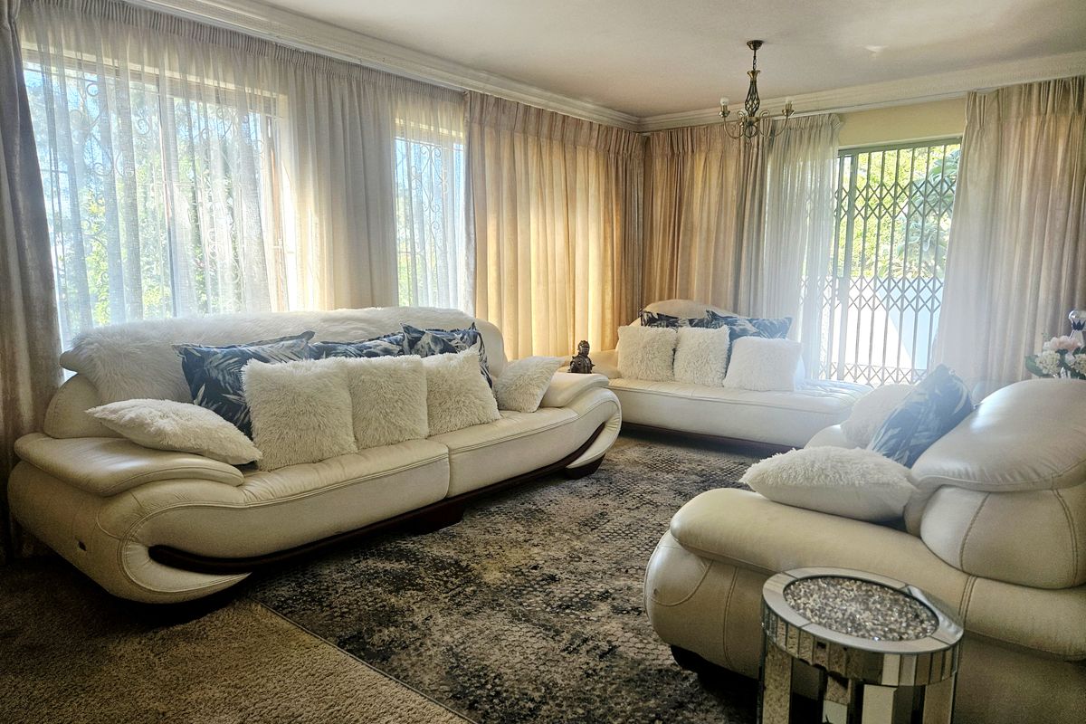 A spacious living room featuring large windows with floor-to-ceiling curtains and sliding glass doors leading to an outdoor area with security gates.