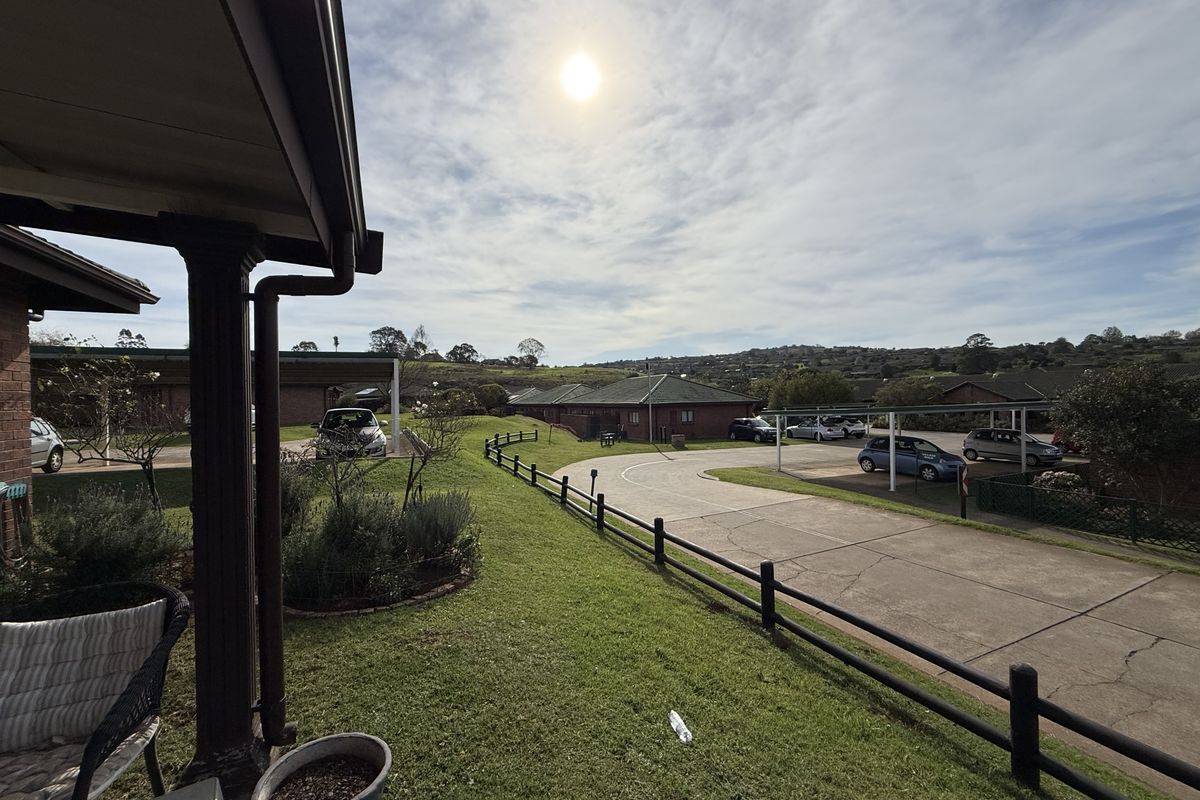 View of the townhouse complex grounds featuring a paved driveway, covered parking bays, and landscaped lawn areas under a bright sky.