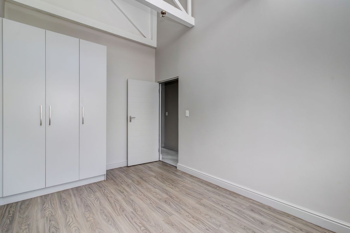 A bright, empty bedroom featuring modern white built-in cupboards, light wood-look laminate flooring, and exposed white ceiling trusses.