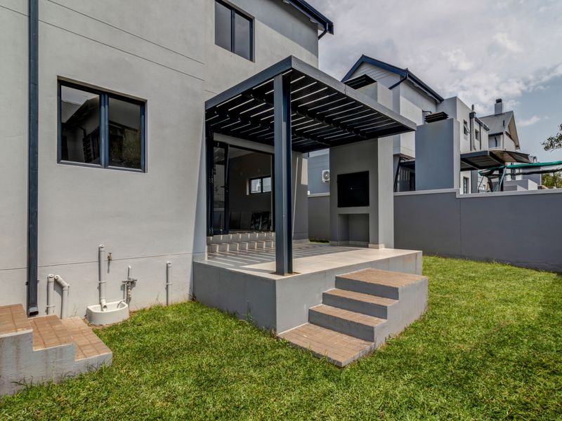 Rear view of the property featuring a covered patio with a built-in braai, concrete steps, and a landscaped garden area.