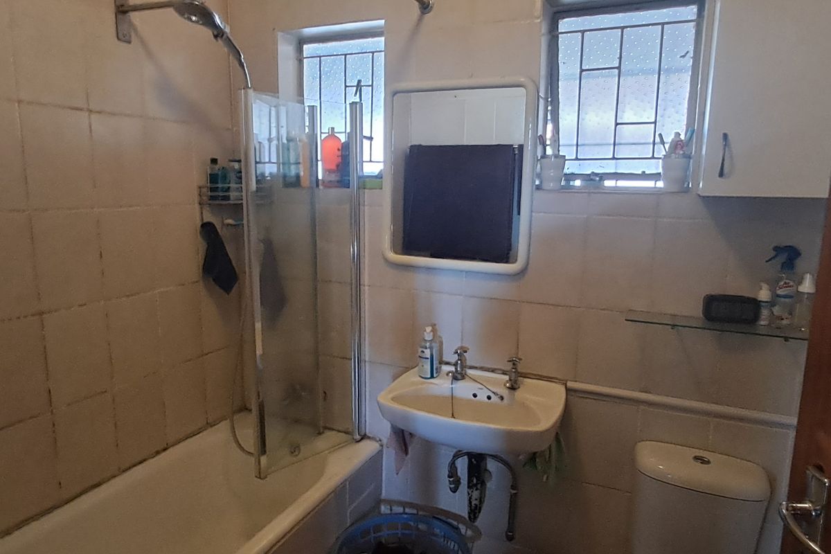 A dated bathroom featuring a bathtub with a shower screen, pedestal sink, toilet, and wall-mounted storage cabinet.