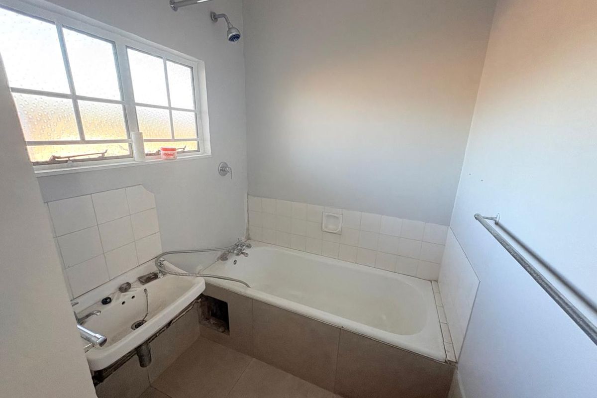 A functional bathroom featuring a built-in bathtub, wall-mounted shower head, and basin, requiring some maintenance and cosmetic updates to the tiling and bath panel.