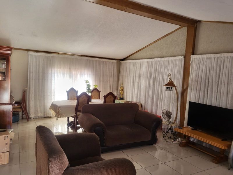 Spacious open-plan living and dining area featuring tiled flooring, a vaulted ceiling with exposed timber beams, and large windows with floor-to-ceiling curtains.