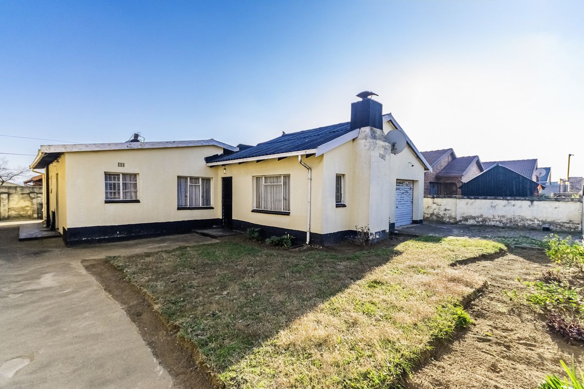 Rear view of a single-story house featuring a chimney, attached garage, and a concrete driveway with an established garden area.