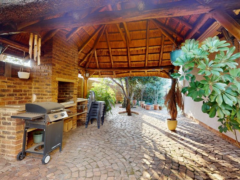 A covered outdoor patio featuring a traditional thatch roof, exposed timber beams, and a built-in brick braai area with cobblestone flooring.