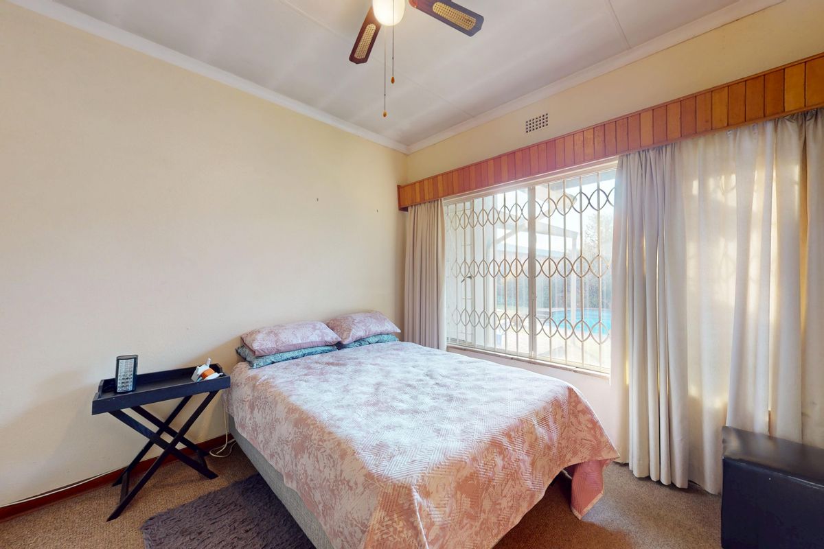 A standard bedroom featuring carpet flooring, a ceiling fan, and a window fitted with a security gate and timber pelmet.