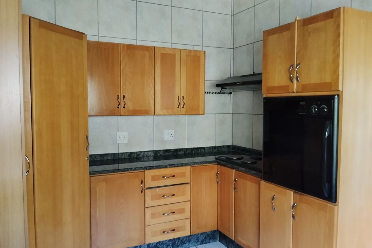 A functional kitchen featuring wooden cabinetry, granite countertops, built-in oven, hob, and extractor fan with neutral tiled splashbacks and flooring.