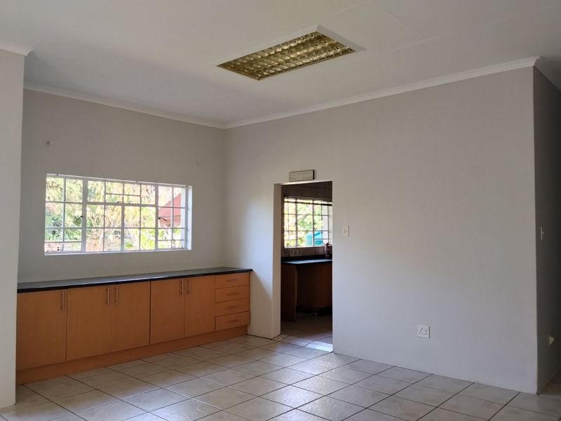 A clean, empty kitchen area featuring built-in wooden cabinetry, dark laminate countertops, tiled flooring, and a large window with security bars.