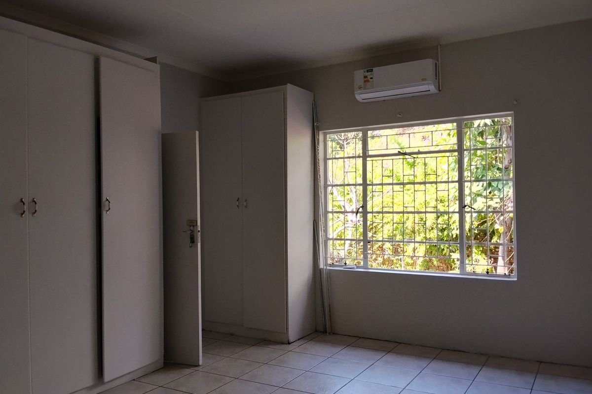 A bright, empty bedroom featuring tiled flooring, built-in cupboards, and a wall-mounted air conditioning unit with security bars on the window.