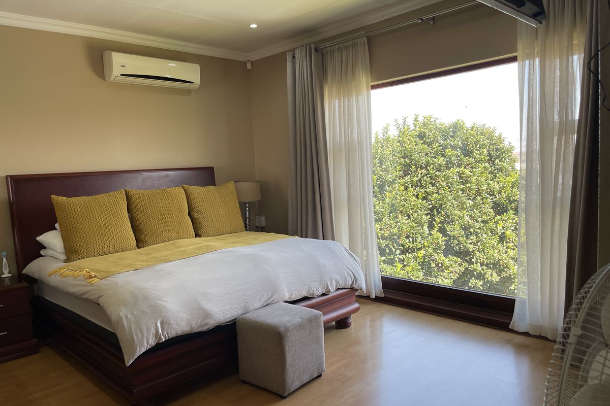 A well-lit bedroom featuring laminate flooring, a wall-mounted air conditioning unit, and a large window providing natural light and garden views.