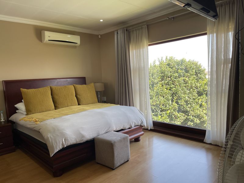A well-lit bedroom featuring laminate flooring, a wall-mounted air conditioning unit, and a large window providing natural light and garden views.