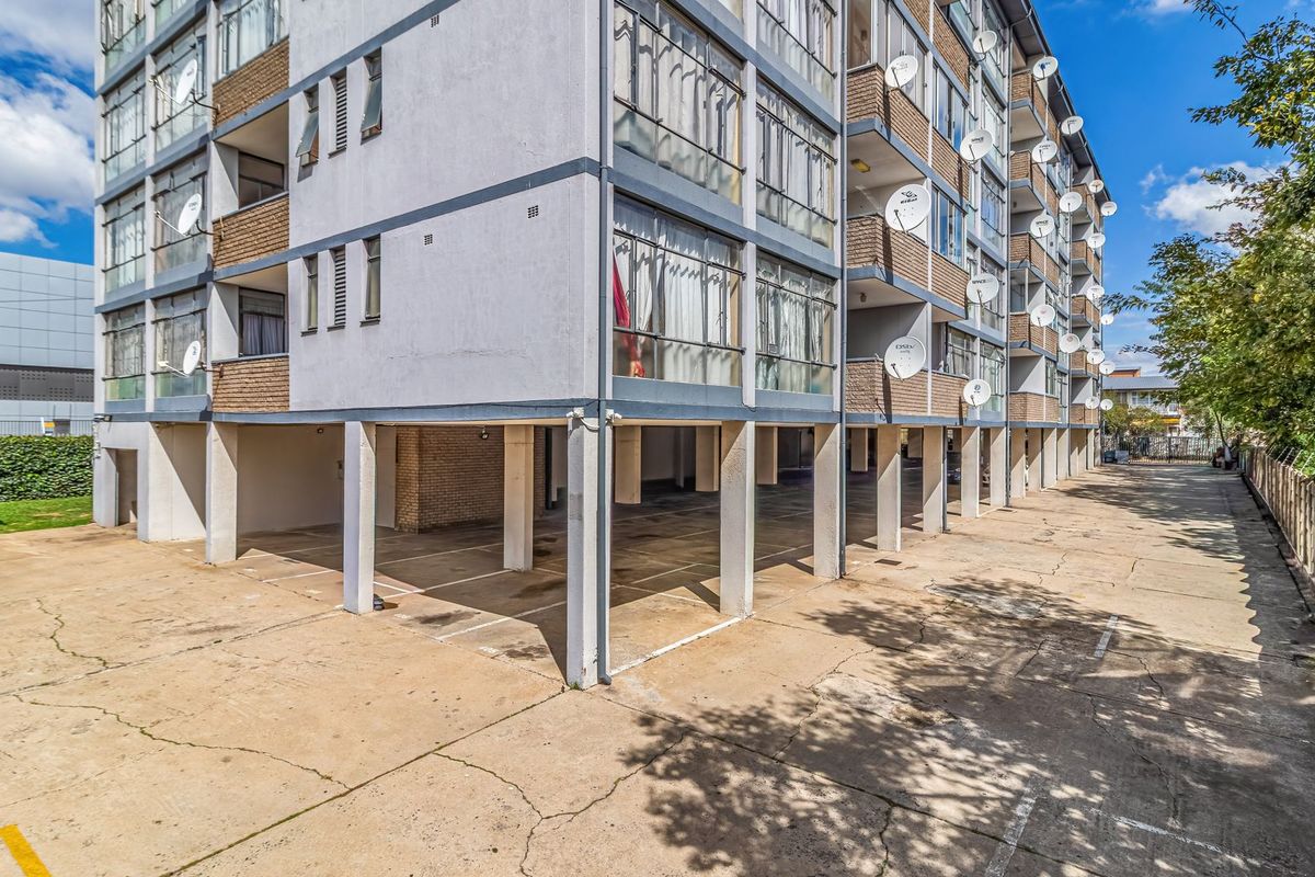 Exterior view of a multi-story apartment building featuring a ground-level covered parking area with concrete pillars and visible satellite dish installations.