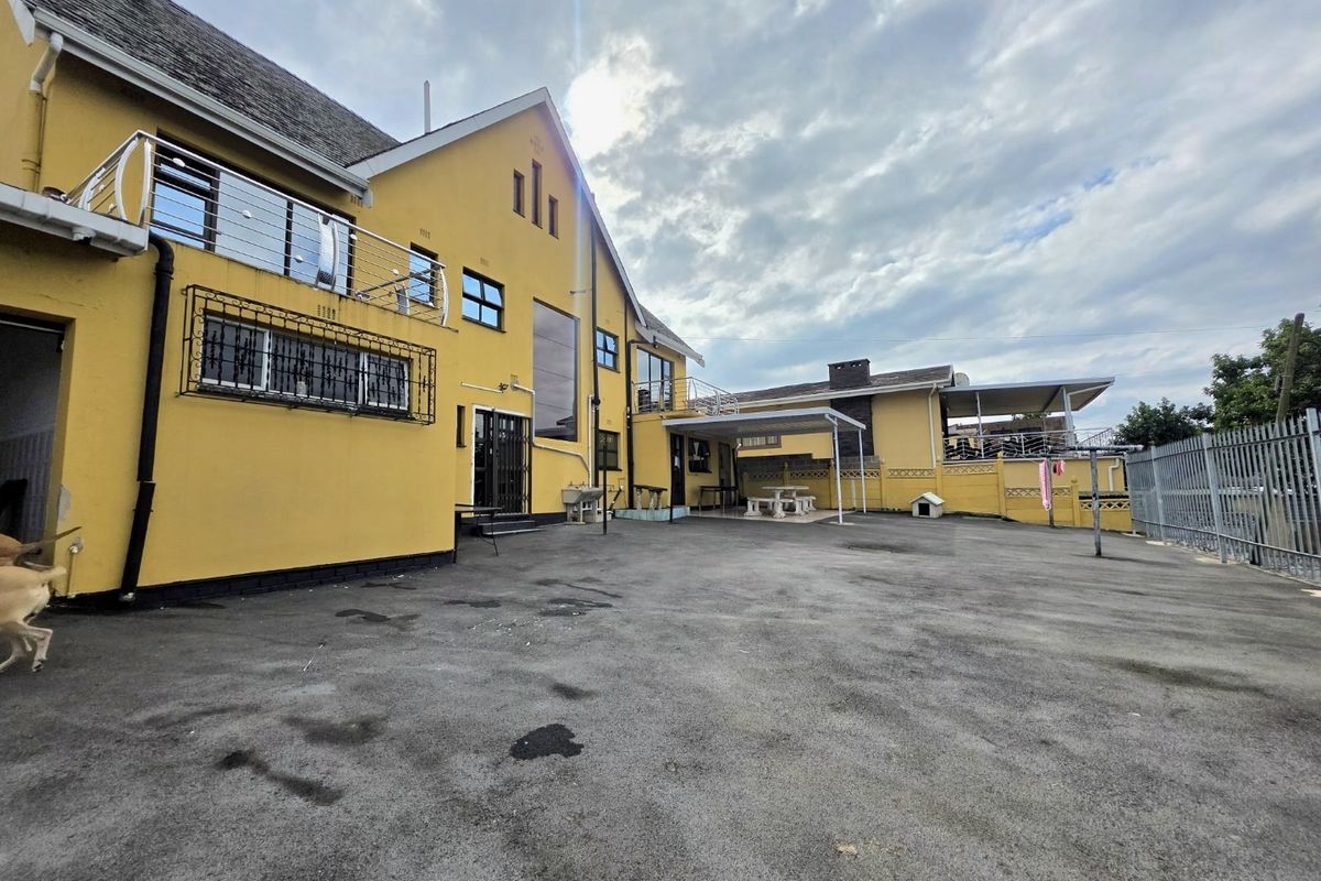 Rear exterior view of the property featuring a large asphalt driveway, covered patio area, and upper-level balcony with stainless steel railings.
