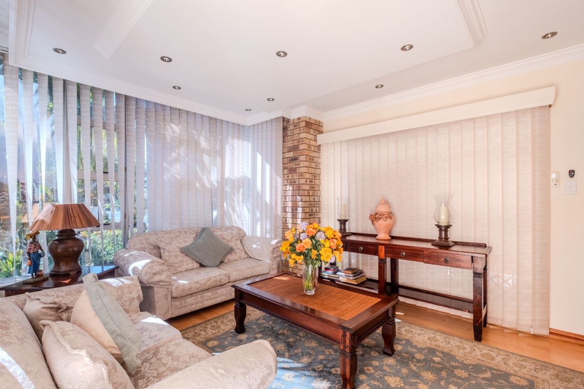 A comfortable living room featuring an exposed brick pillar, recessed downlighting, cove ceiling detailing, and large windows with vertical blinds.