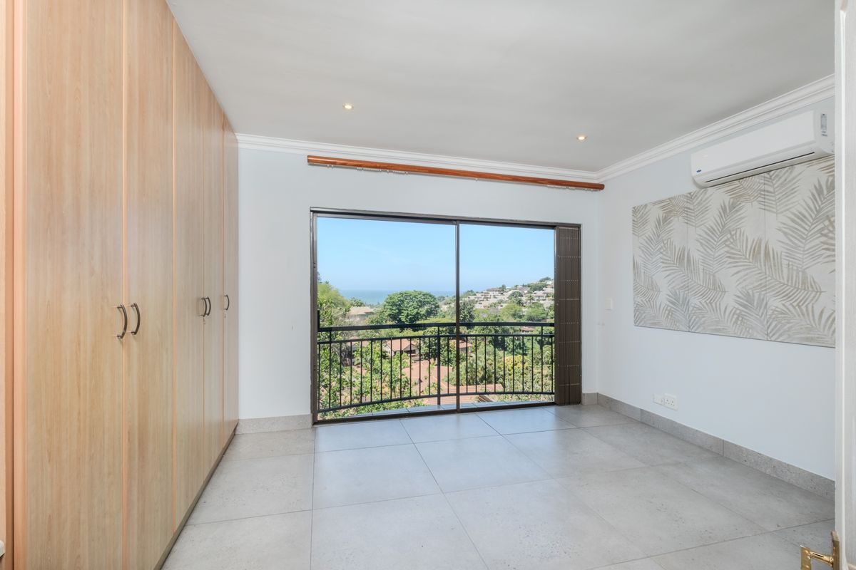 A bright, empty bedroom featuring built-in cupboards, tiled flooring, air conditioning, and a sliding door opening to a balcony with scenic views.