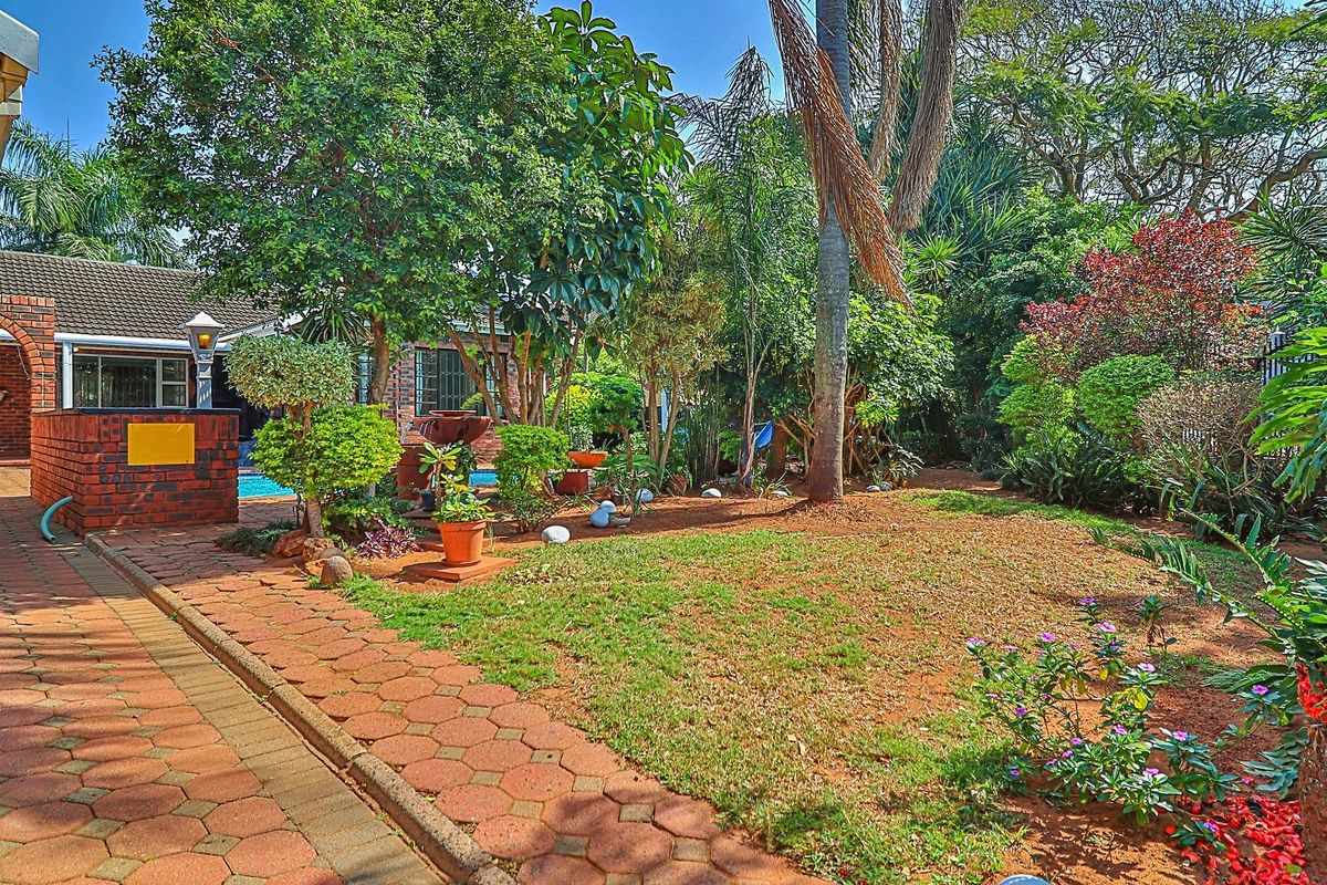 A well-maintained rear garden featuring brick paving, mature trees, and a glimpse of the swimming pool area.