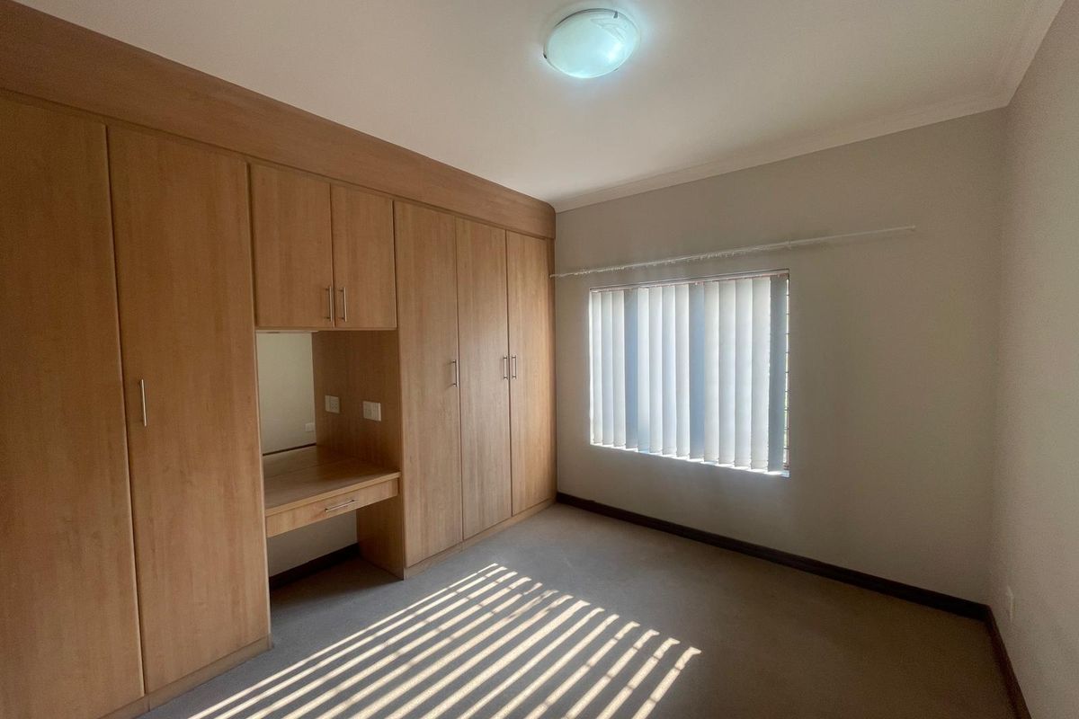 A bright, empty bedroom featuring extensive built-in wooden cupboards with an integrated vanity desk, carpeted flooring, and vertical window blinds.