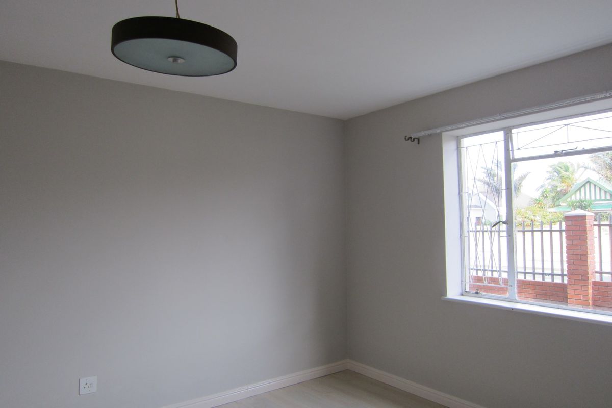 A clean, neutral-toned bedroom featuring a window with security bars, a pendant light fixture, and light-colored flooring.