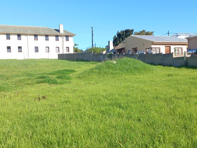 A view of a vacant residential plot featuring a grass lawn and concrete boundary wall, surrounded by neighboring residential buildings.