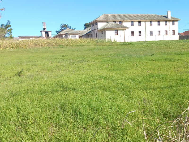 A view of a vacant residential plot of land with grass cover, showing adjacent buildings in the background.