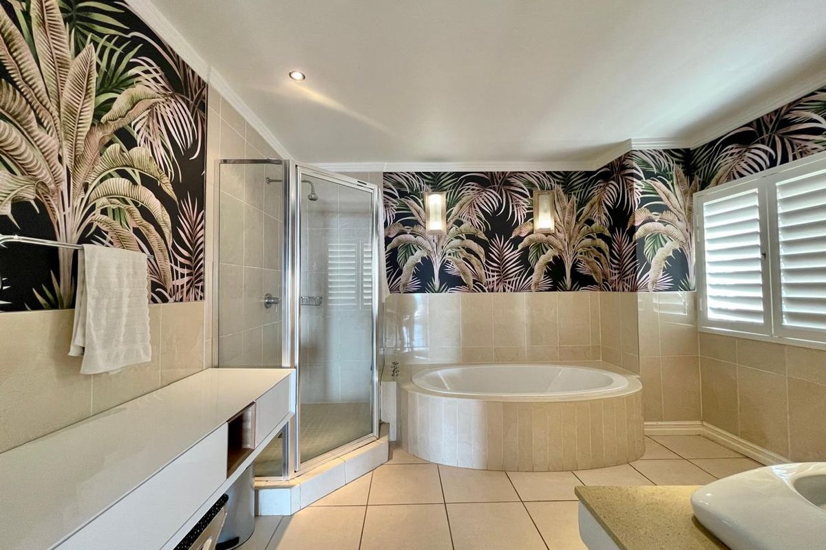 Spacious bathroom featuring a built-in corner bathtub, separate shower enclosure, vanity unit, and decorative wallpaper with integrated lighting and shutter blinds.