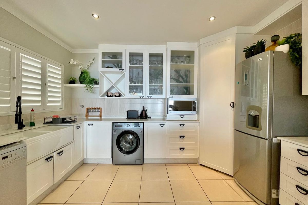 A well-maintained kitchen featuring white cabinetry, a farmhouse sink, glass-fronted display units, and integrated appliances with neutral tiled flooring.