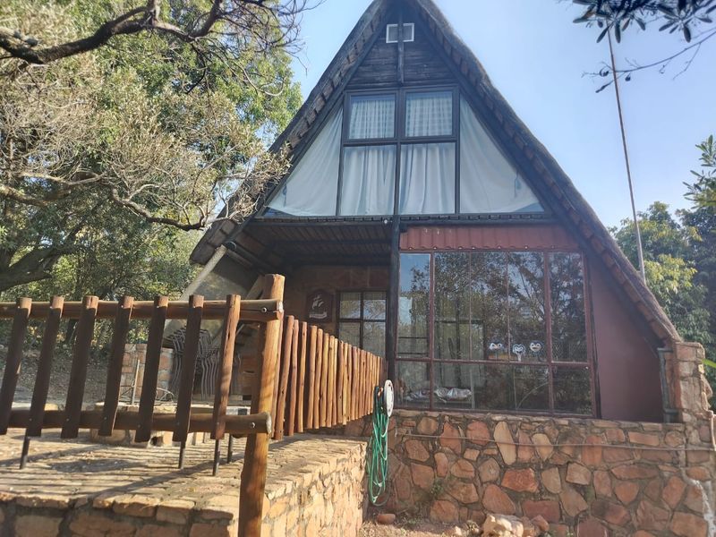 A rustic A-frame house featuring stone cladding, a timber deck with balustrade, and large glass windows set in a natural, wooded environment.