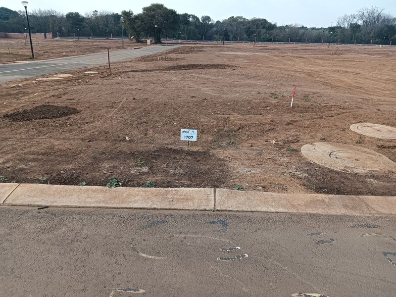 A vacant residential plot featuring a cleared surface, paved road access, concrete curbing, and established utility access points.