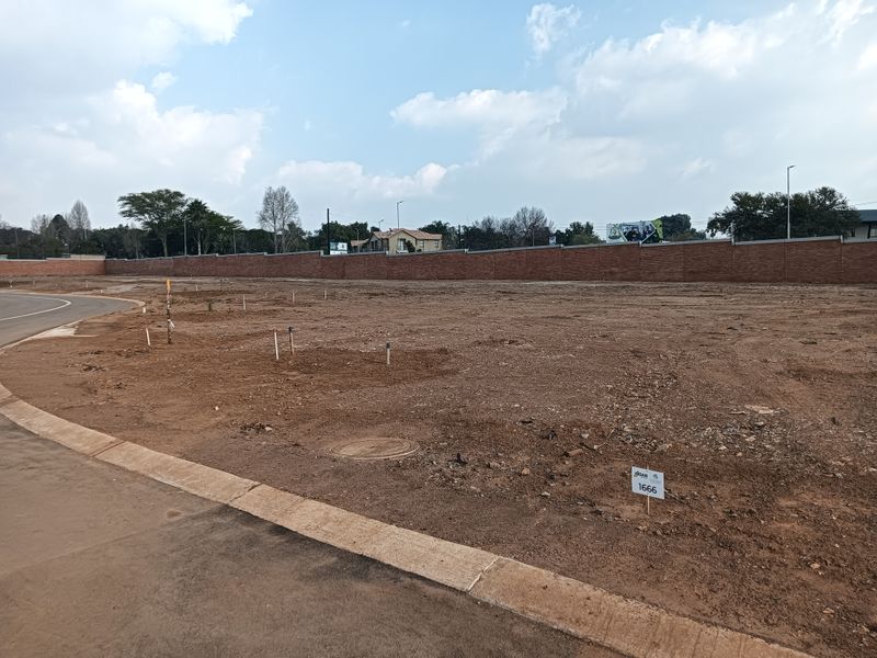 A vacant residential plot featuring a paved road frontage, survey markers, and a secure brick perimeter wall.