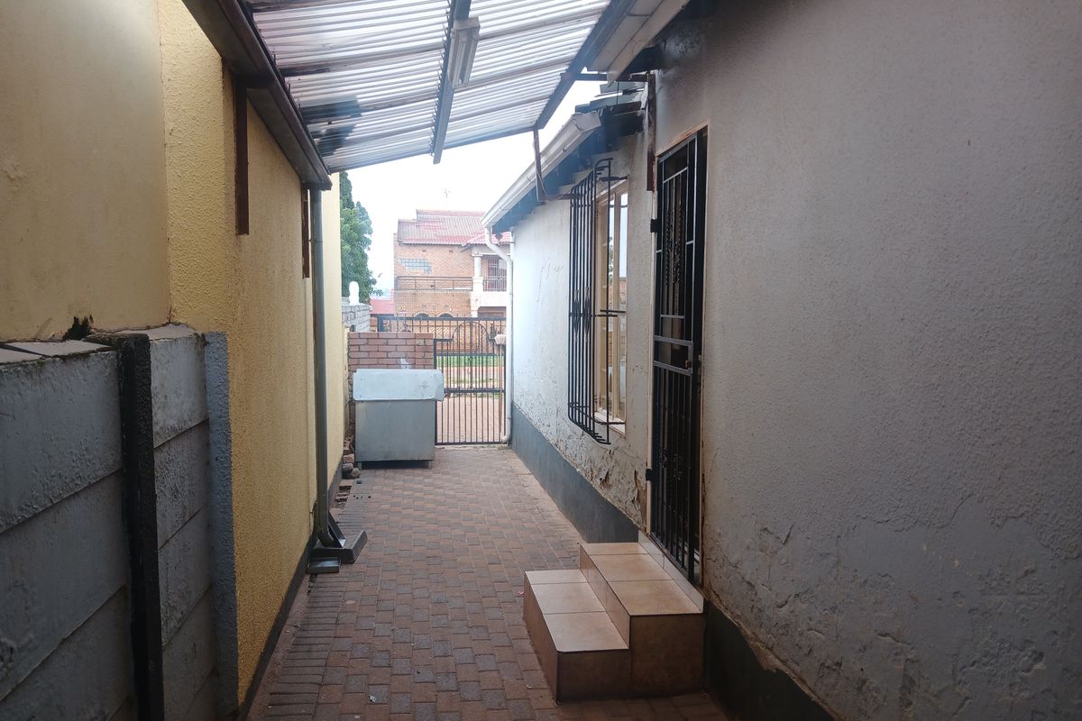 A narrow side passage featuring paved flooring, a covered walkway, and security fixtures, showing signs of exterior wear and weathering.