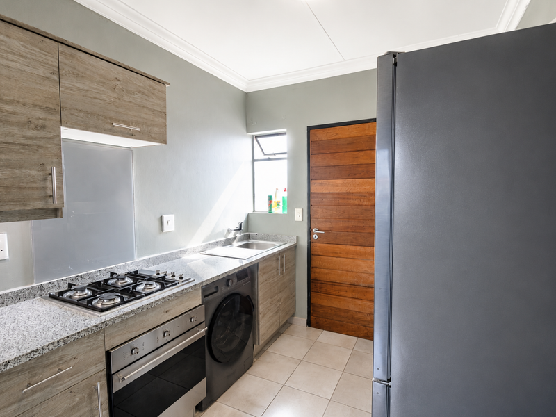 Modern kitchen featuring wood-finish cabinetry, granite countertops, integrated gas hob, built-in oven, and tiled flooring with direct access to an exterior door.