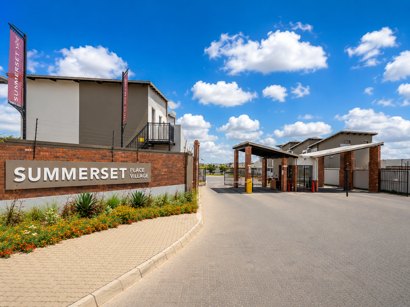 The entrance to Summerset Place Village featuring a brick boundary wall, paved driveway, security gate, and guard house.