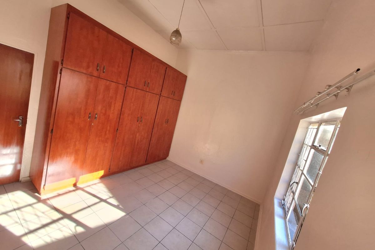 A vacant bedroom featuring tiled flooring, built-in wooden cupboards, and a window fitted with security bars.