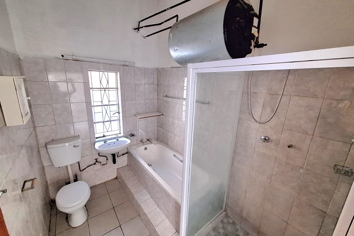 A functional bathroom featuring a built-in bathtub, separate shower enclosure, pedestal basin, and toilet, with exposed geyser and dated tiling.