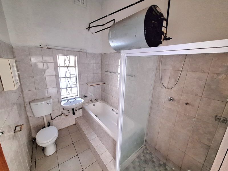 A functional bathroom featuring a built-in bathtub, separate shower enclosure, pedestal basin, and toilet, with exposed geyser and dated tiling.
