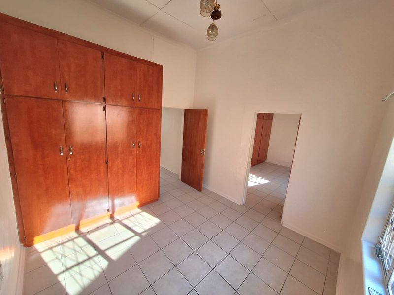 A spacious, empty bedroom featuring built-in wooden cupboards, tiled flooring, and a view into an adjacent room.