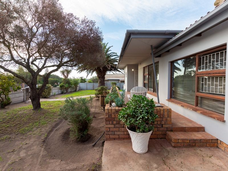 A rear exterior view featuring a covered patio with brick steps, overlooking a garden area with established trees and perimeter fencing.