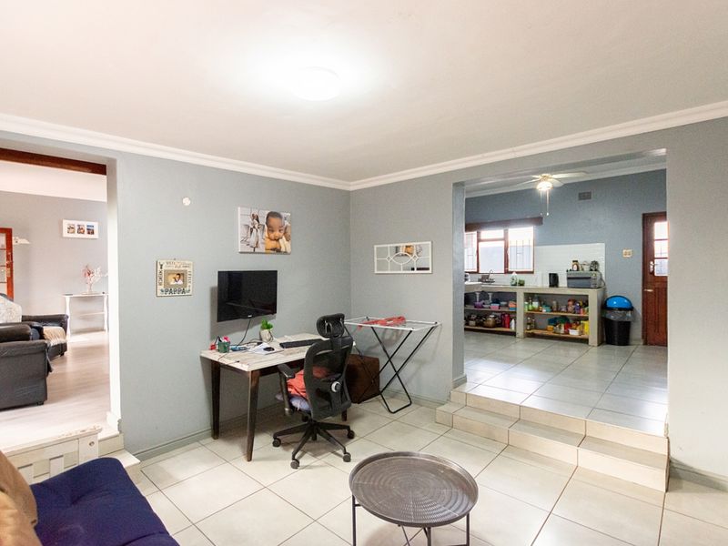 An open-plan living area featuring tiled flooring, a raised kitchen transition, and neutral wall tones, connecting to adjacent living and kitchen spaces.