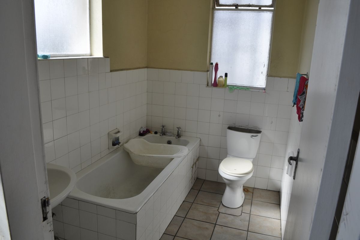 A dated bathroom featuring a tiled bathtub, pedestal basin, and toilet, with significant wear to the wall and floor finishes.