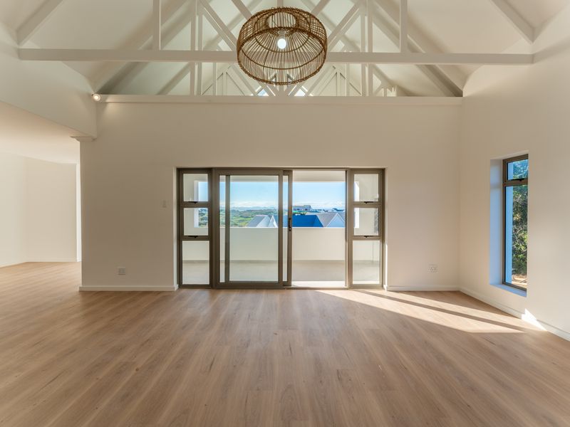 Spacious open-plan living area featuring high vaulted ceilings with exposed trusses, light wood-look flooring, and large sliding doors opening to a balcony with views.
