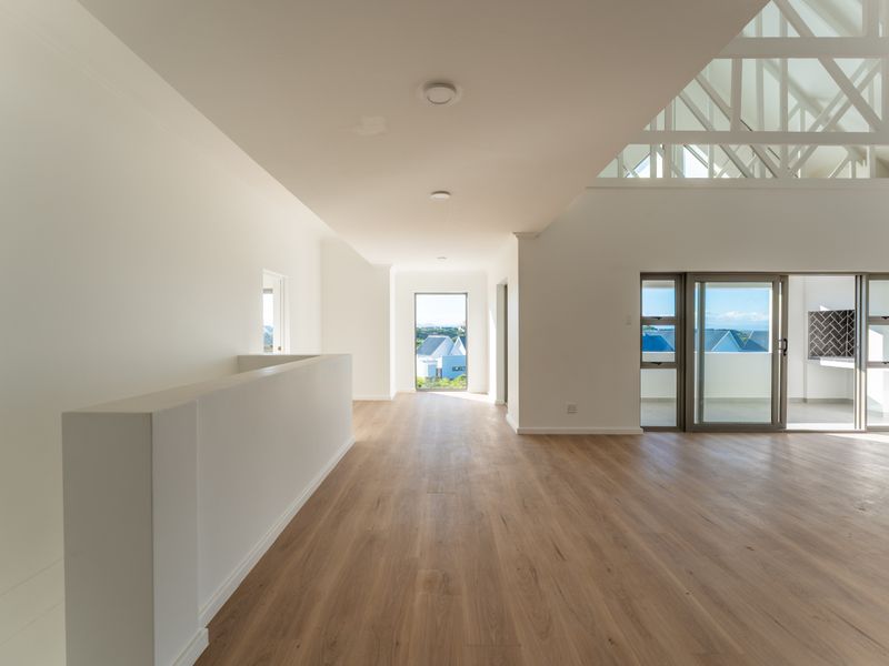 Spacious open-plan living area featuring high vaulted ceilings with exposed trusses, light wood-look flooring, and large sliding glass doors leading to a balcony.