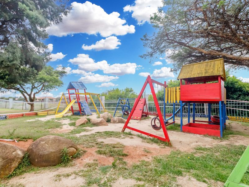 A communal outdoor garden area featuring a playground with various climbing structures, swings, and metal perimeter fencing.