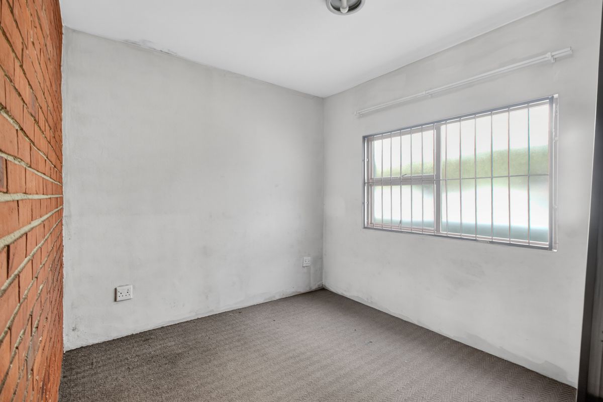 An empty bedroom featuring an exposed brick feature wall, carpeted flooring, and a window fitted with security bars.