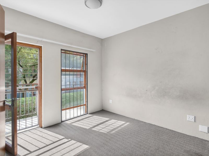 An empty bedroom featuring carpet flooring, security bars on windows, and direct access to a balcony, with some visible wear on the walls.