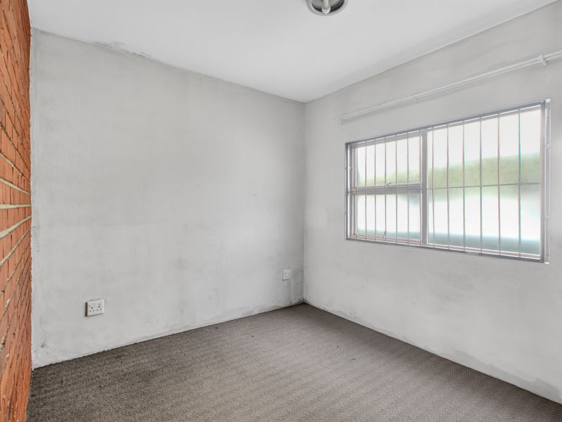 An empty bedroom featuring an exposed brick feature wall, carpeted flooring, and a window fitted with security bars.