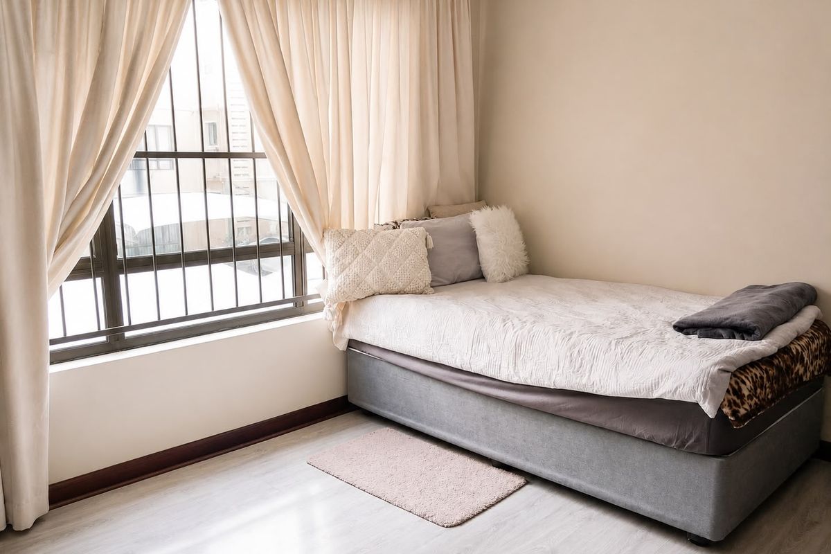 A bright bedroom featuring light-toned laminate flooring, neutral walls, and a large window fitted with security bars.